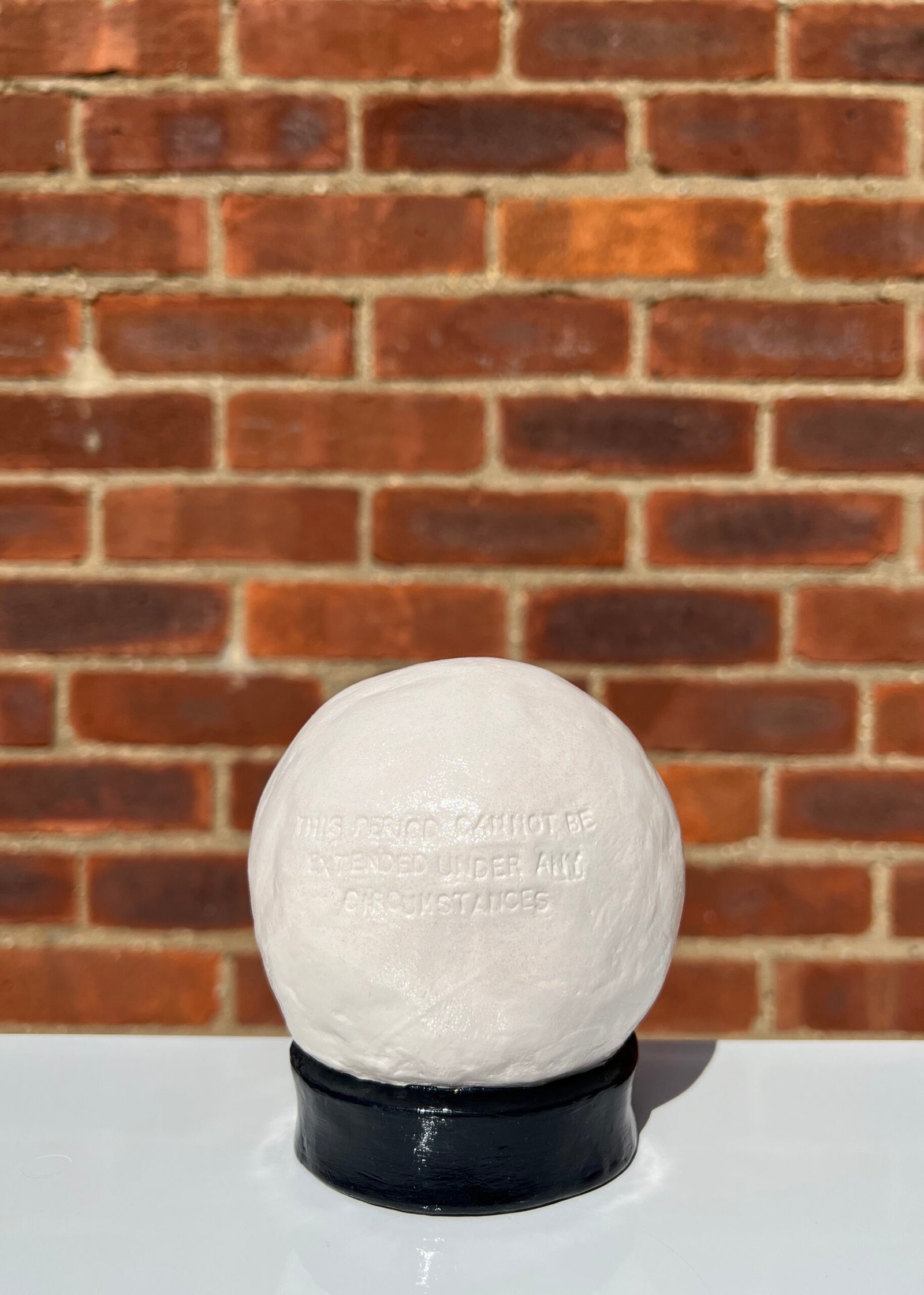 A handmade clay crystal ball blind-embossed with the uppercase text "this period cannot be extended under any circumstances". The ball is painted off-white and coated in clear varnish with very fine iridescent glitter. The ball is attached to a round-collared base painted glossy black. The photograph is taken outdoors in direct sunlight against the backdrop of a brick wall. The crystal ball is sat on a white surface casting a shadow behind it.