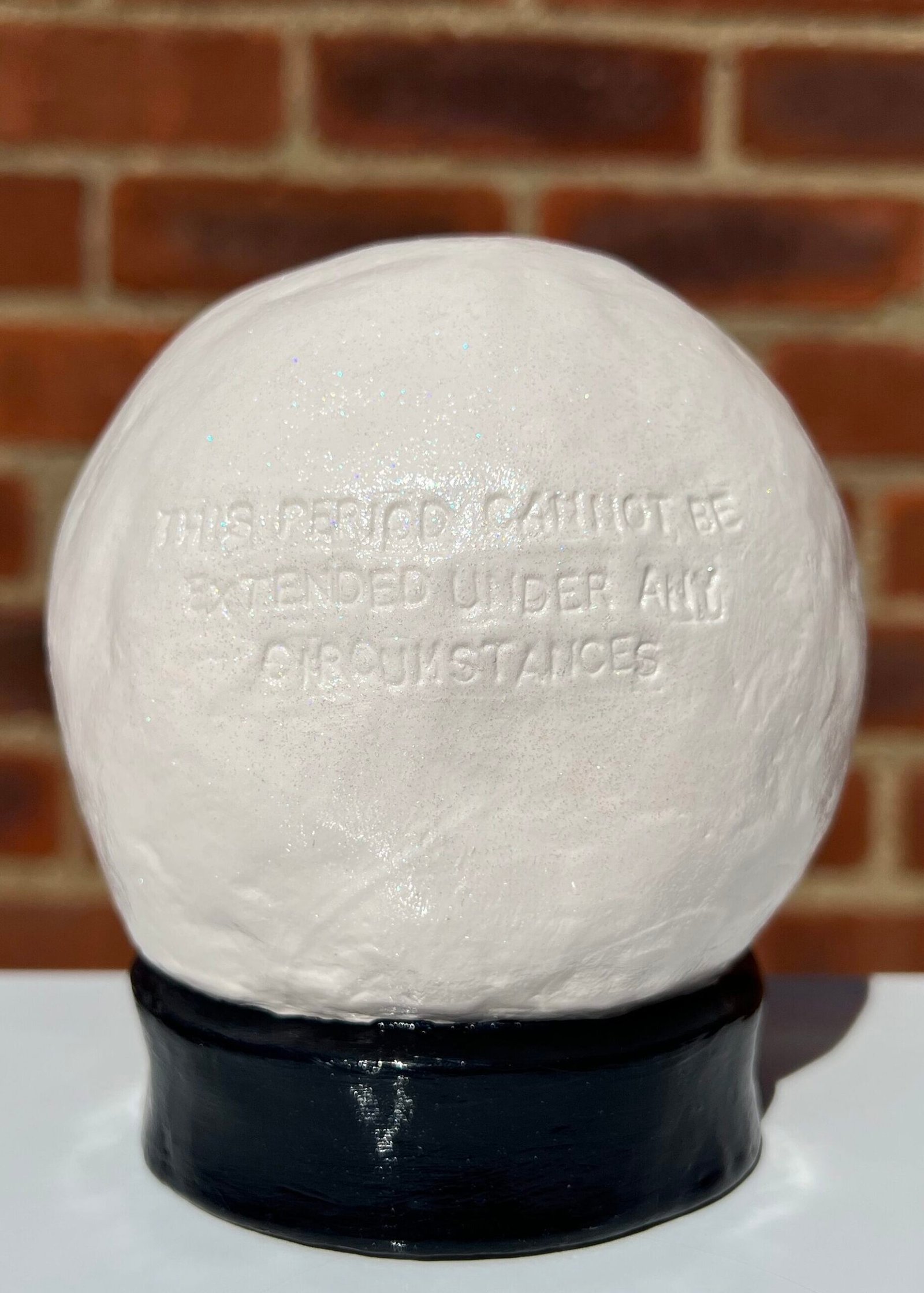 A handmade clay crystal ball blind-embossed with the uppercase text "this period cannot be extended under any circumstances". The ball is painted off-white and coated in clear varnish with very fine iridescent glitter. The ball is attached to a round-collared base painted glossy black. The photograph is taken outdoors in direct sunlight against the backdrop of a brick wall. The crystal ball is sat on a white surface casting a shadow behind it.