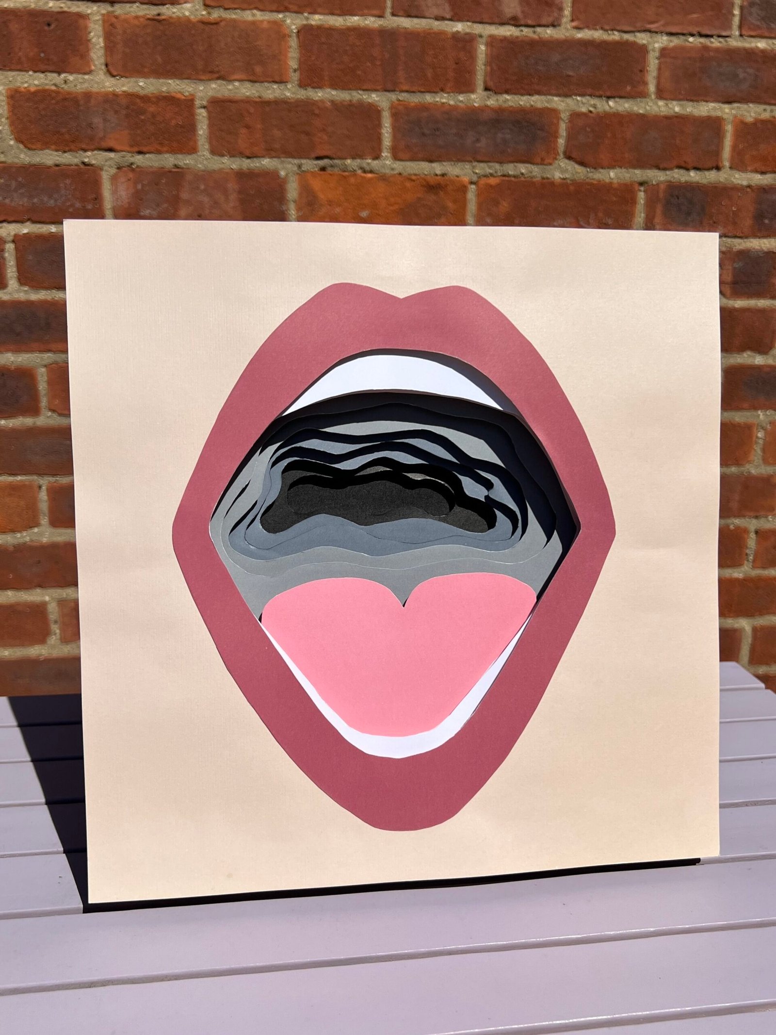 A multi-layered paper-cutting in a graphic style depicting a wide open mouth. Bordered by a beige, square background, a dark pink, wide-open mouth frames rows of white teeth and a pale pink heart-shaped tongue. The back of the throat is grey, cut out with an unruly, wavy edge. Each layer beneath descends deeper into an increasingly dark, chaotic, and tight void, until reaching a black hole. The photograph is taken outdoors in direct sunlight against the backdrop of a brick wall. The piece is sat on a lilac slatted table casting a shadow behind it.