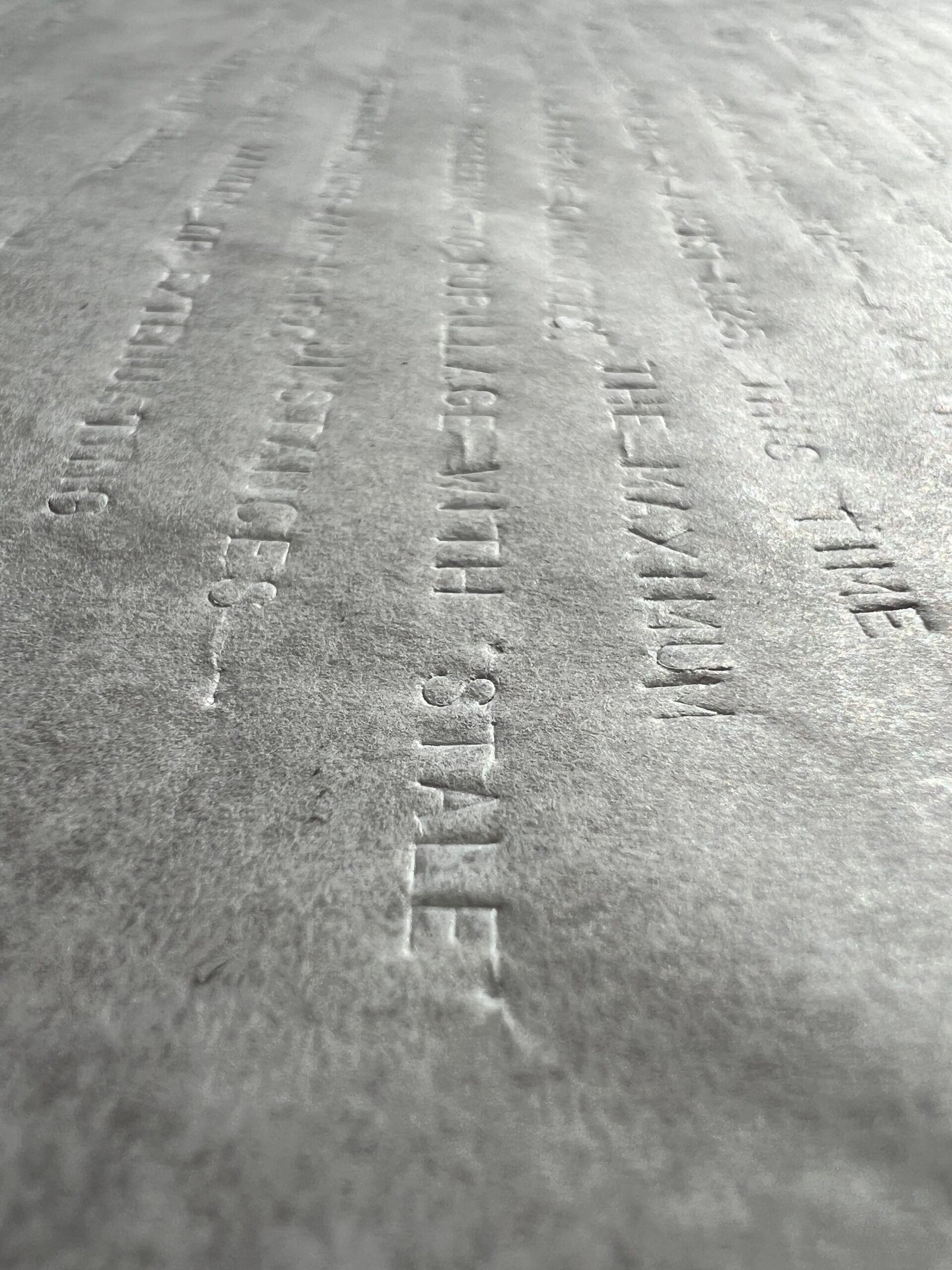 A close-up photograph of a large sheet of parchment paper which has been blind debossed with uppercase text in a font similar used by typewriters. The translucency and lack of ink make the debossed text visible but illegible except at specific angles, under certain light, and on close inspection. The photograph and lighting are angled enabling the words to be deciphered. The words 'TIME', 'MAXIMUM' and 'STALE' stand out.